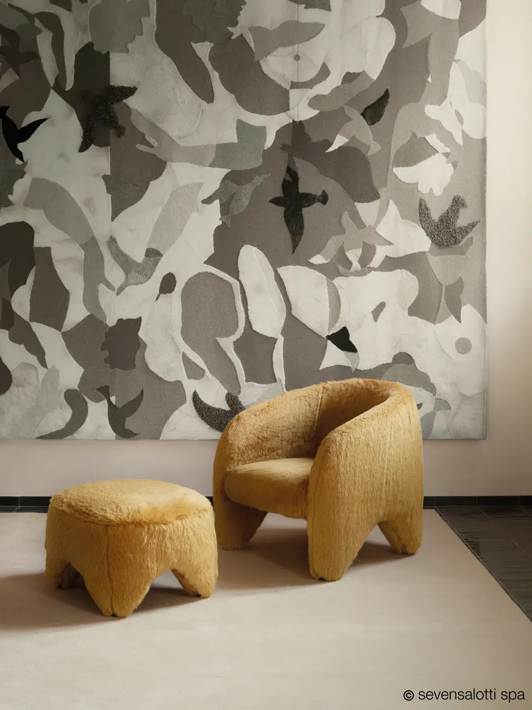 Poltrona Yuzu Arflex a pelo lungo color ocra con pouf davanti a un grande arazzo grigio